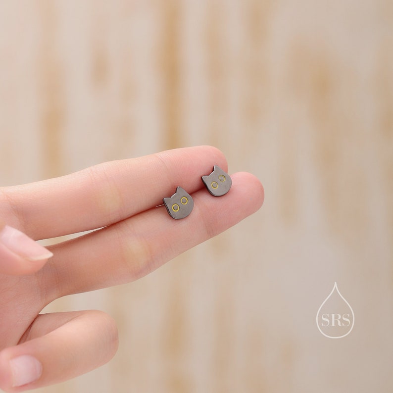 Peut inclure: Une paire de boucles d'oreilles clous en forme de chat noir avec des accents dor&eacute;s. Les boucles d'oreilles sont montr&eacute;es sur un doigt.