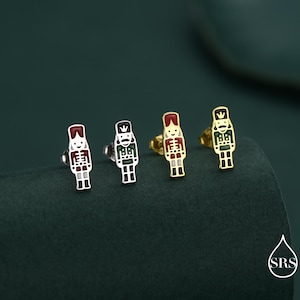 Puede incluir: Cuatro pendientes de botón con un diseño de soldado cascanueces colorido. Los pendientes son en tonos plateados y dorados, con detalles de esmalte rojo, verde y blanco. Se muestran sobre una superficie verde oscuro.