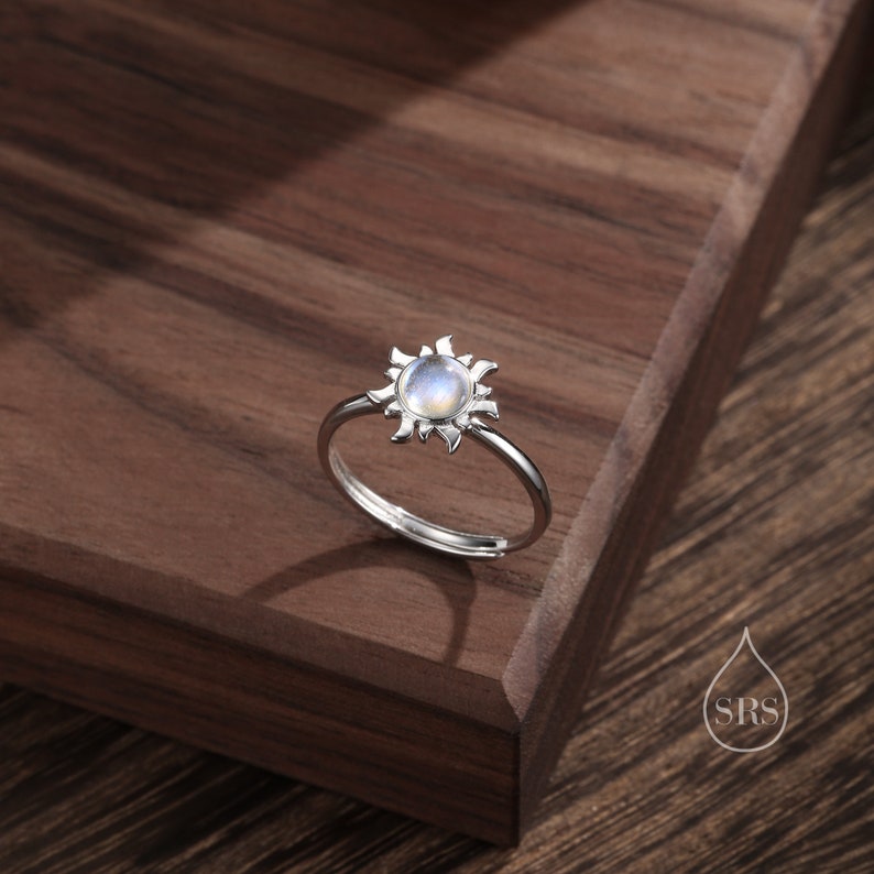 Puede incluir: Un anillo de plata con una piedra de luna engastada en un dise&ntilde;o de sol. El anillo est&aacute; sobre una superficie de madera.