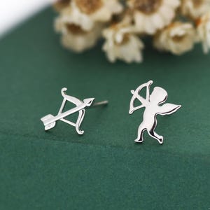 May include: A pair of silver stud earrings. One earring shows a bow and arrow, the other a small cupid figure with wings. The earrings are set against a green background with blurred white flowers.