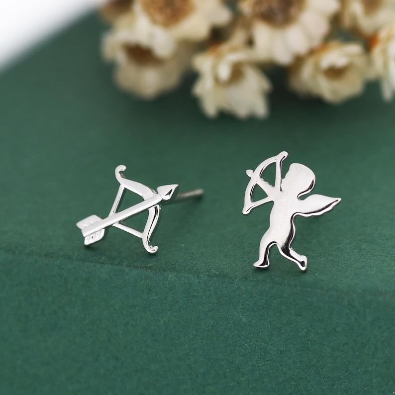 May include: A pair of silver stud earrings. One earring shows a bow and arrow, the other a small cupid figure with wings. The earrings are set against a green background with blurred white flowers.