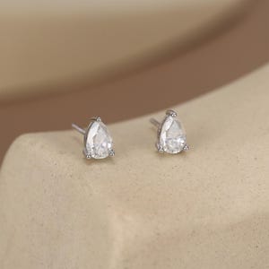 May include: A pair of silver stud earrings, each featuring a pear-shaped, clear gemstone. The earrings are set on a neutral-toned surface, showcasing their sparkling facets. The studs are small and delicate, perfect for everyday wear.
