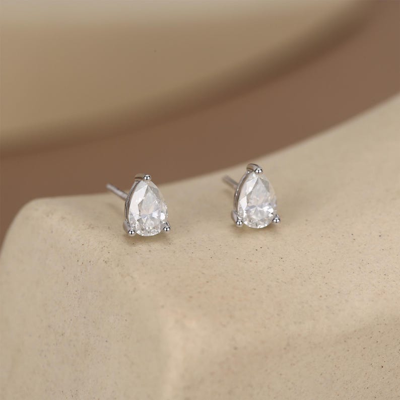 May include: A pair of silver stud earrings, each featuring a pear-shaped, clear gemstone. The earrings are set on a neutral-toned surface, showcasing their sparkling facets. The studs are small and delicate, perfect for everyday wear.