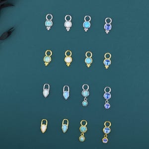 Può includere: Una collezione di piccoli ciondoli decorativi per gioielli in tonalità argento e oro. I ciondoli presentano pietre preziose opalescenti e blu di varie forme e dimensioni. I ciondoli sono disposti su uno sfondo verde scuro.