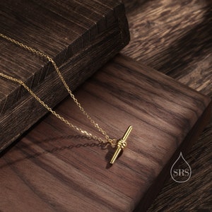 May include: A gold necklace with a delicate chain and a simple bar pendant with a knot detail. The necklace is displayed on a wooden surface.