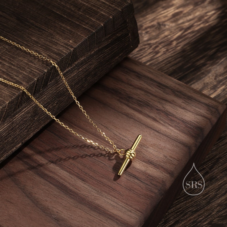 May include: A gold necklace with a delicate chain and a simple bar pendant with a knot detail. The necklace is displayed on a wooden surface.
