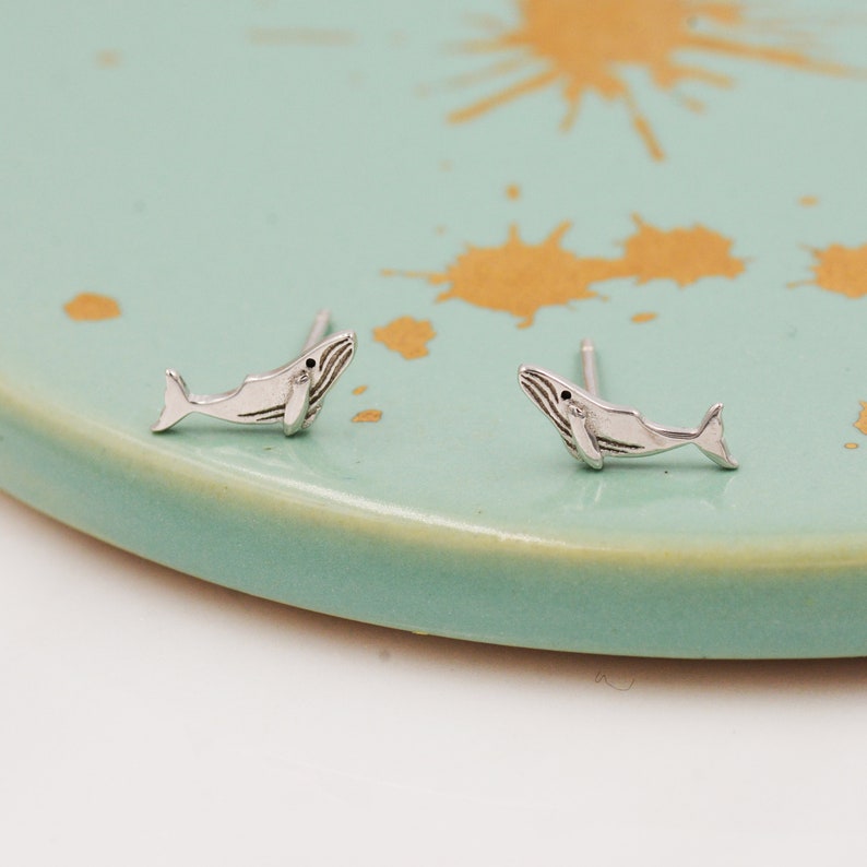 May include: A pair of silver whale stud earrings on a light blue background.