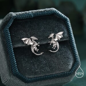 May include: A pair of silver dragon stud earrings. Each earring features a detailed dragon with outstretched wings, set against a dark teal velvet jewelry box. The earrings are small and delicate.