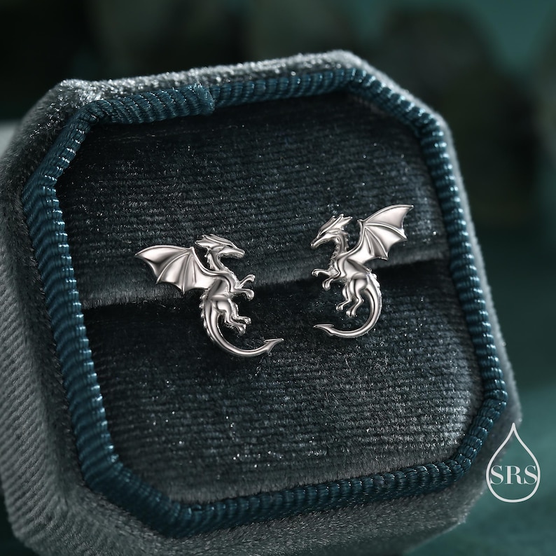 May include: A pair of silver dragon stud earrings. Each earring features a detailed dragon with outstretched wings, set against a dark teal velvet jewelry box. The earrings are small and delicate.