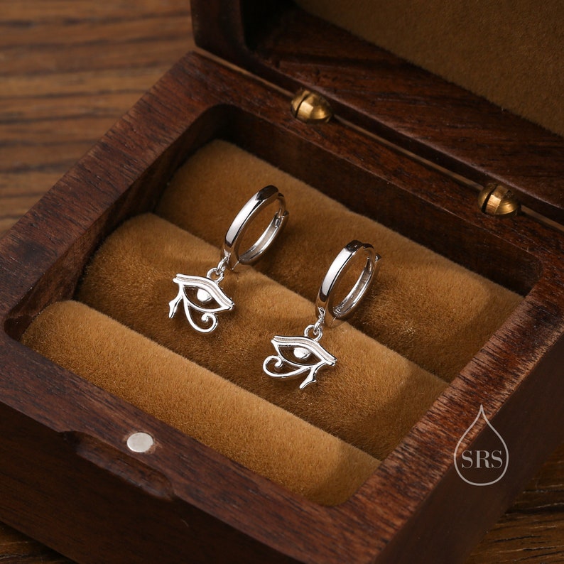 May include: A pair of silver hoop earrings with a dangling eye of Horus charm. The earrings are displayed in a wooden box.