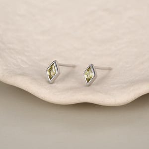 Puede incluir: Un par de pendientes de botón con una piedra preciosa en forma de cometa de color verde oliva claro, engastada en un marco de metal plateado pulido. Cada pendiente tiene un poste recto y fino en la parte posterior para el lóbulo.