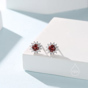 May include: A pair of silver stud earrings with a red gemstone surrounded by smaller white gemstones.