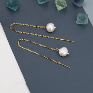 May include: A pair of gold chain earrings with a white pearl dangling from each end. The earrings are on a dark blue surface.