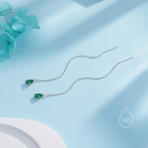 May include: A pair of silver threader earrings with teardrop-shaped, emerald green gemstones. The earrings hang from delicate chains, creating a graceful, flowing design. The background is a soft, light blue, with a touch of floral decoration.