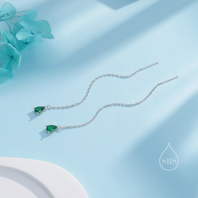 May include: A pair of silver threader earrings with teardrop-shaped, emerald green gemstones. The earrings hang from delicate chains, creating a graceful, flowing design. The background is a soft, light blue, with a touch of floral decoration.
