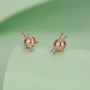 Pode incluir: Um par de brincos de ouro rosa. Cada brinco apresenta uma bola de l&atilde; com tr&ecirc;s agulhas de tric&ocirc;. Os brincos est&atilde;o sobre um fundo verde claro.
