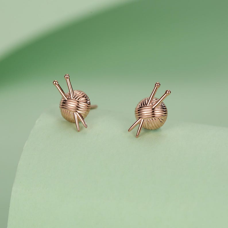 Pode incluir: Um par de brincos de ouro rosa. Cada brinco apresenta uma bola de l&atilde; com tr&ecirc;s agulhas de tric&ocirc;. Os brincos est&atilde;o sobre um fundo verde claro.