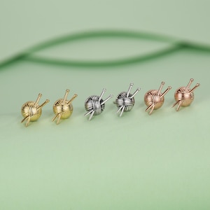 Pode incluir: Seis brincos de bot&atilde;o pequenos em fila, cada um representando uma bola de l&atilde; com agulhas de tric&ocirc;. Os brincos s&atilde;o em tons de ouro, prata e ouro rosa, em um fundo verde suave.