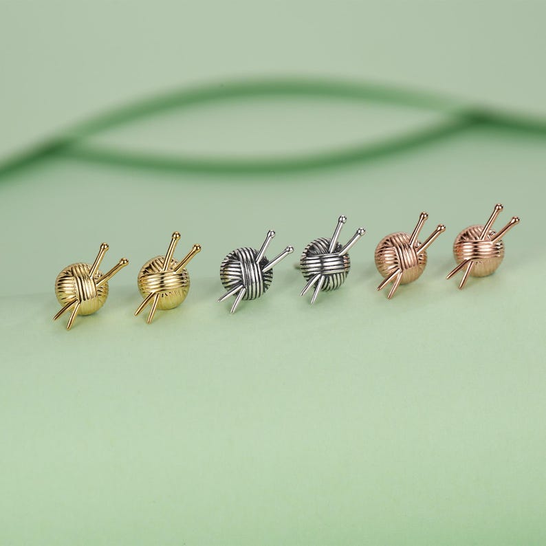 Pode incluir: Seis brincos de bot&atilde;o pequenos em fila, cada um representando uma bola de l&atilde; com agulhas de tric&ocirc;. Os brincos s&atilde;o em tons de ouro, prata e ouro rosa, em um fundo verde suave.