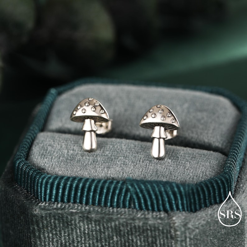 May include: A pair of silver mushroom stud earrings. Each earring features a mushroom cap with small, evenly spaced dots and a stem. The earrings are displayed in a teal velvet jewelry box, with a blurred green background.