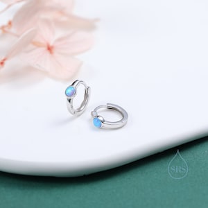 Puede incluir: Un par de pendientes de aro de plata con una pequeña piedra de ópalo azul en cada pendiente.