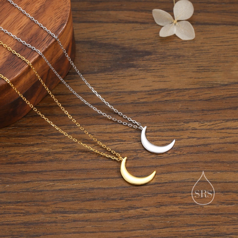 May include: Two delicate crescent moon necklaces on a wooden surface. One necklace is gold and the other is silver.