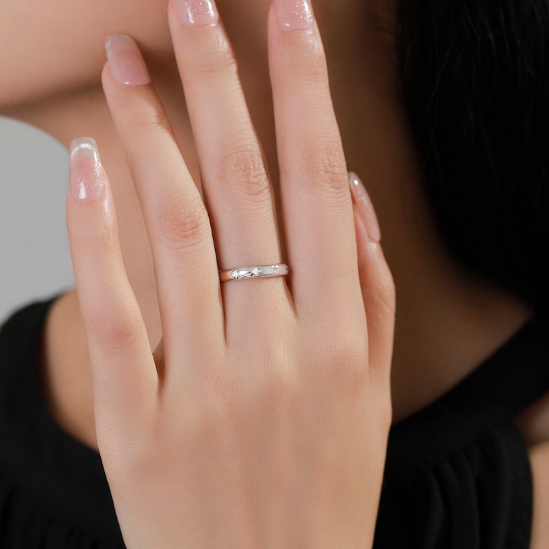 May include: A thin silver band ring with a textured surface. The ring is worn on the ring finger of a person's hand.