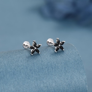 May include: Two silver-tone flower stud earrings with black gemstone petals. The earrings are displayed on a blue surface. Each earring has a post for pierced ears.