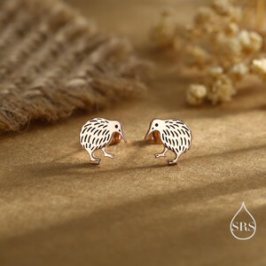 May include: A pair of rose gold kiwi bird stud earrings. Each earring features a detailed kiwi design with a textured body and a long beak. The earrings are displayed on a textured, golden-brown surface.