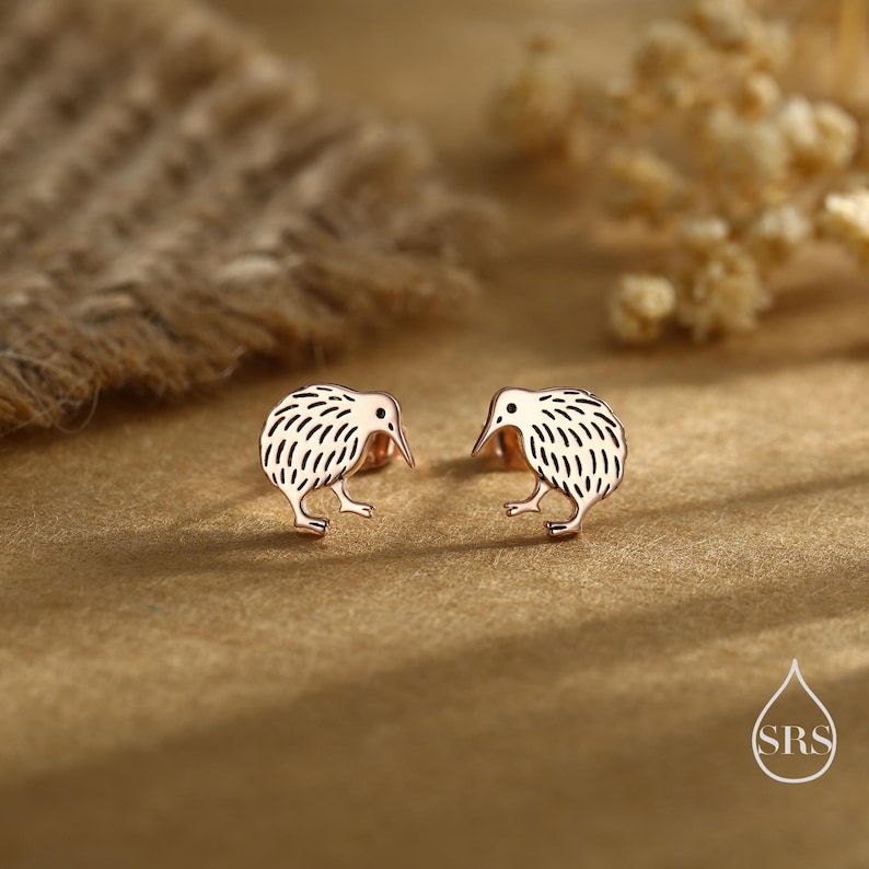 May include: A pair of rose gold kiwi bird stud earrings. Each earring features a detailed kiwi design with a textured body and a long beak. The earrings are displayed on a textured, golden-brown surface.