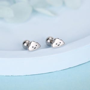 May include: A pair of silver cloud-shaped stud earrings with a smiling face. The earrings have a post and ball backing. The earrings are displayed on a light blue surface.