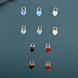 Op de afbeelding: Een verzameling hartvormige bedels in verschillende kleuren en afwerkingen. Sommige bedels zijn zilver- of goudkleurig met iriserende, opalen, zwarte of rode emaille harten. Elke bedel heeft een kleine lus aan de bovenkant om te bevestigen.