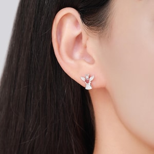 May include: A pair of silver angel stud earrings. Each earring features an angel design with a red heart-shaped center. The angel has wings and a halo. The earrings are small and delicate.