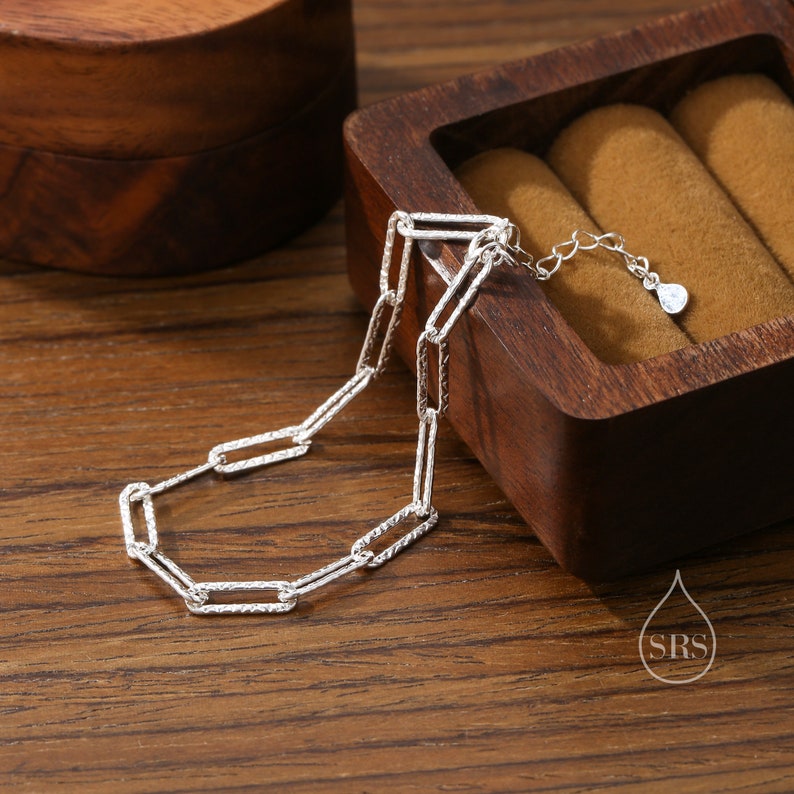 May include: A silver chain bracelet with a textured, rectangular link design. The bracelet is lying on a wooden surface.