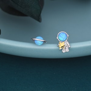 May include: A pair of silver-toned stud earrings. One earring is a blue planet with a silver ring, and the other is an astronaut holding a gold star. The astronaut's helmet and the planet are blue.