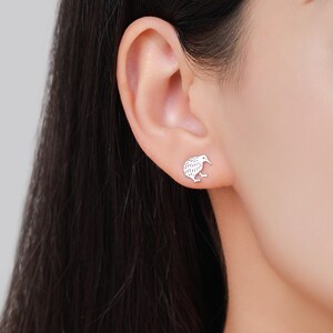 May include: A pair of silver-colored stud earrings, each shaped like a kiwi bird. The earrings are small and detailed, with the bird's features clearly visible. The earrings are shown on an ear, suggesting they are being worn.