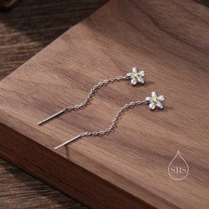 May include: A pair of silver dangle earrings with a small white flower charm and a gold center. The earrings have a delicate chain that hangs down from the flower.
