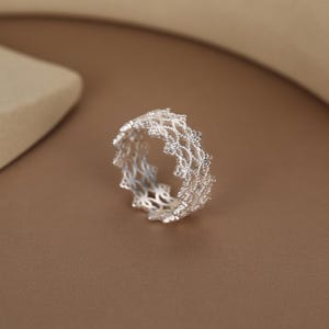 May include: A silver ring featuring an intricate, openwork lace-like pattern. The band is constructed from multiple rows of small, interconnected metallic loops that create a delicate, textured, and airy appearance throughout the entire circular structure.