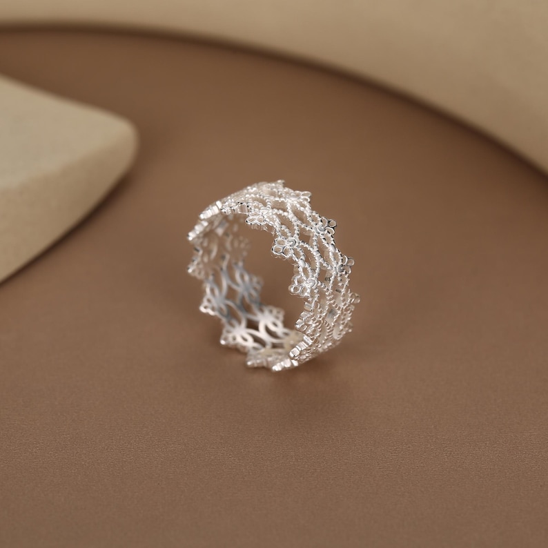 May include: A silver ring featuring an intricate, openwork lace-like pattern. The band is constructed from multiple rows of small, interconnected metallic loops that create a delicate, textured, and airy appearance throughout the entire circular structure.
