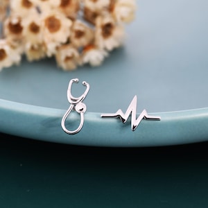 Pode incluir: Brincos de pressão prateados com estetoscópio e batimento cardíaco. Os brincos estão sobre uma superfície azul clara com flores brancas e amarelas desfocadas no fundo. O brinco de estetoscópio é uma miniatura detalhada.