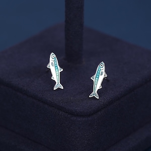 Può includere: Un paio di orecchini a bottone a forma di pesce argentato. Ogni orecchino presenta un corpo bianco con dettagli blu e un design dettagliato. Sono esposti su una superficie blu scuro.