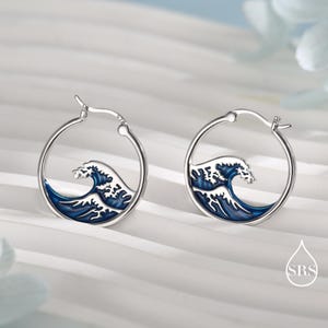 Puede incluir: Un par de pendientes de aro de plata con un dise&ntilde;o circular y una incrustaci&oacute;n de esmalte que representa una ola oce&aacute;nica estilizada en azul y blanco. Los pendientes tienen un mecanismo de cierre de bisagra y un acabado met&aacute;lico pulido.