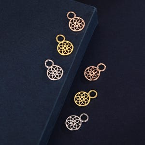 Pode incluir: Seis pingentes metálicos circulares com um padrão floral geométrico vazado. O conjunto inclui dois pingentes em ouro rosa, dois em ouro amarelo e dois em tom prateado, cada um com um pequeno anel circular no topo para fixação.