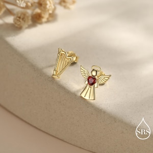 May include: A pair of gold-colored stud earrings. One earring is shaped like a harp, and the other is an angel with a red heart-shaped gem. The angel has wings and a halo. The earrings are on a neutral-colored surface.