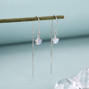 May include: A pair of silver threader earrings. Each earring features a small, iridescent star charm suspended from a delicate chain. The earrings are displayed against a light blue background, with a wooden stick in the background.