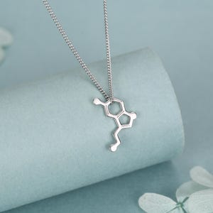 May include: A silver-toned necklace featuring a pendant shaped like a chemical molecular structure. The pendant is suspended from a delicate, thin silver-toned chain. The design consists of interconnected hexagonal and pentagonal geometric shapes.