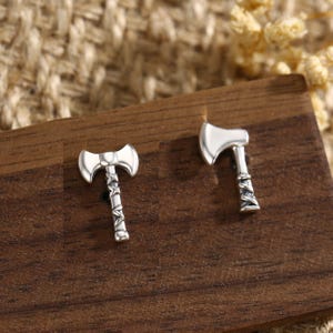 May include: A pair of silver axe-shaped stud earrings. The earrings are presented on a dark wooden surface. The axe heads are smooth and shiny, while the handles have a detailed, textured design.