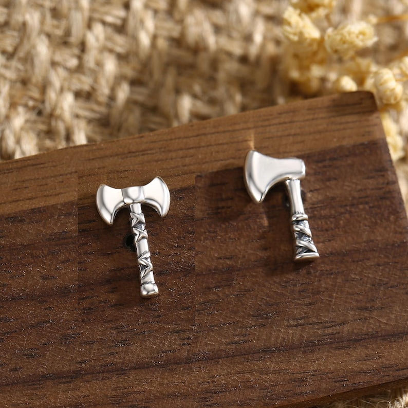 May include: A pair of silver-colored axe-shaped stud earrings. The earrings are displayed on a dark wooden surface. The axe heads are smooth and shiny, while the handles have a detailed, textured design.