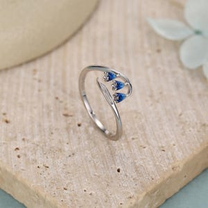 Pu&ograve; includere: Un anello d'argento con un design floreale, caratterizzato da tre boccioli di fiori blu e una foglia. L'anello &egrave; presentato su uno sfondo neutro, evidenziando i delicati dettagli del gioiello.