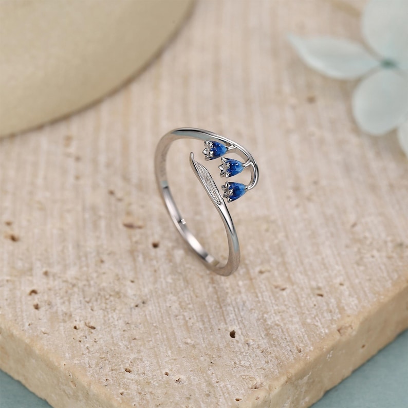Pu&ograve; includere: Un anello d'argento con un design floreale, caratterizzato da tre boccioli di fiori blu e una foglia. L'anello &egrave; presentato su uno sfondo neutro, evidenziando i delicati dettagli del gioiello.